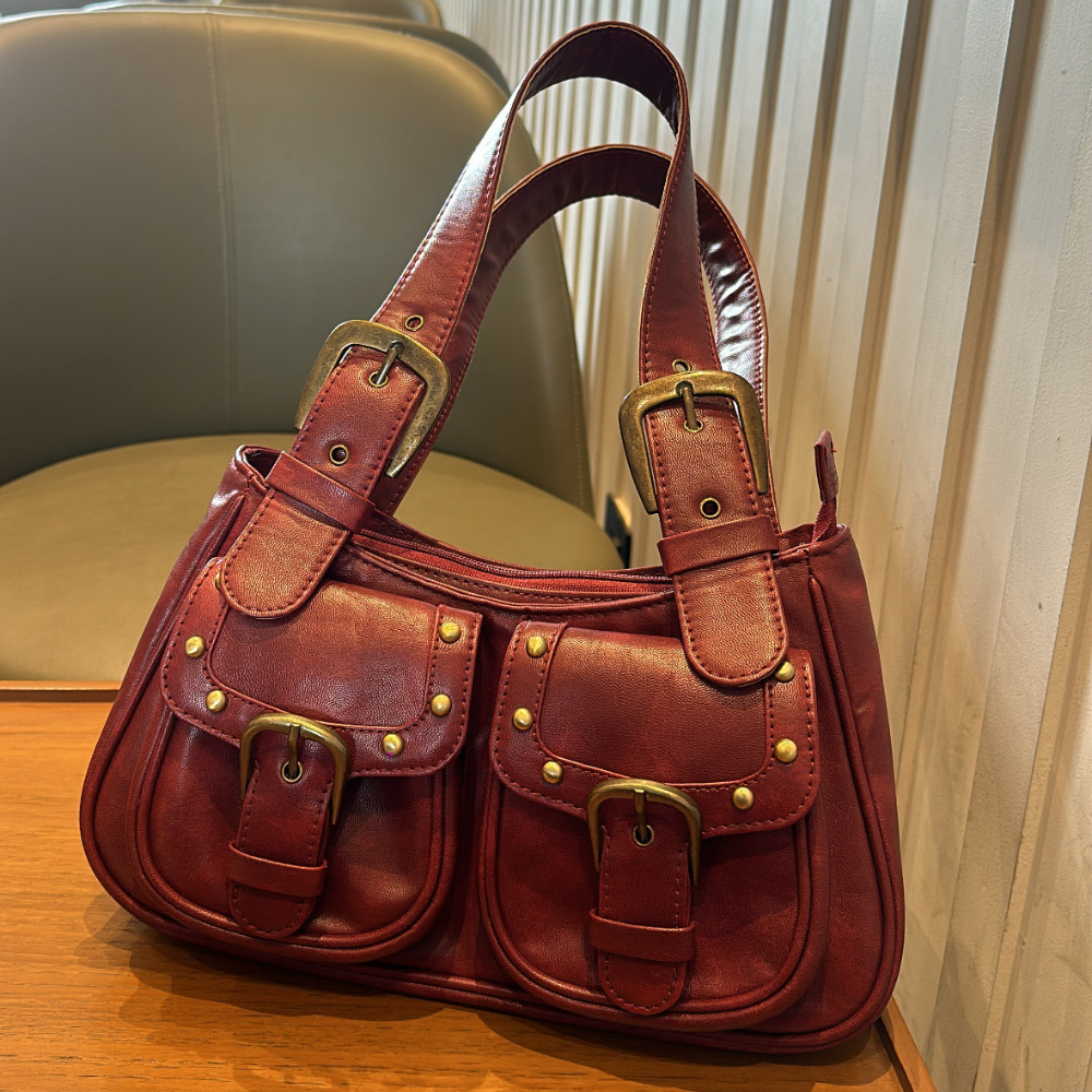 A vintage-inspired red leather satchel with brass-tone buckles, stud detailing, and dual shoulder straps, hanging against a textured white background.