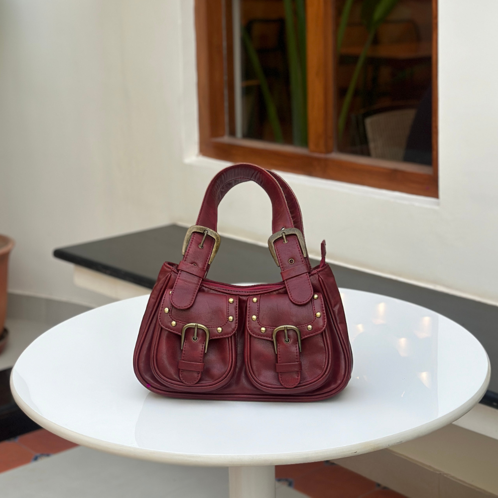 A vintage-inspired red leather satchel with brass-tone buckles, stud detailing, and dual shoulder straps, hanging against a textured white background.