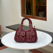 A vintage-inspired red leather satchel with brass-tone buckles, stud detailing, and dual shoulder straps, hanging against a textured white background.